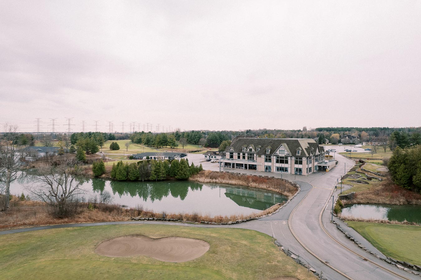Aerial view of a picturesque golf course with a clubhouse beside a serene lake, surrounded by lush greenery and trees, under an overcast sky.