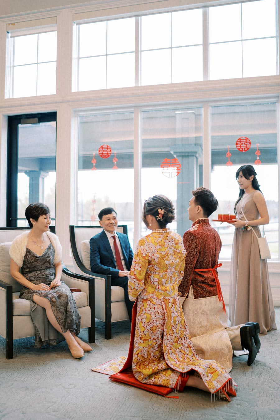 A traditional Vietnamese tea ceremony taking place indoors with a couple in vibrant cultural attire kneeling before seated elders, accompanied by a woman serving tea, against a backdrop of large windows adorned with red decorations.