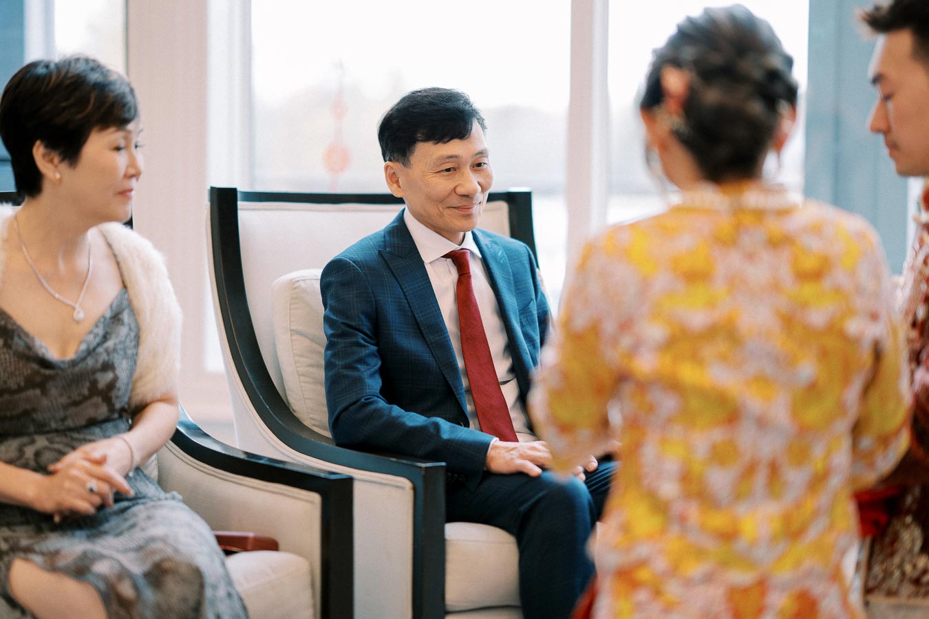 A group of people conversing in a formal setting, with a man in a blue suit and red tie sitting on a chair, surrounded by others dressed elegantly, possibly at a family gathering or event.