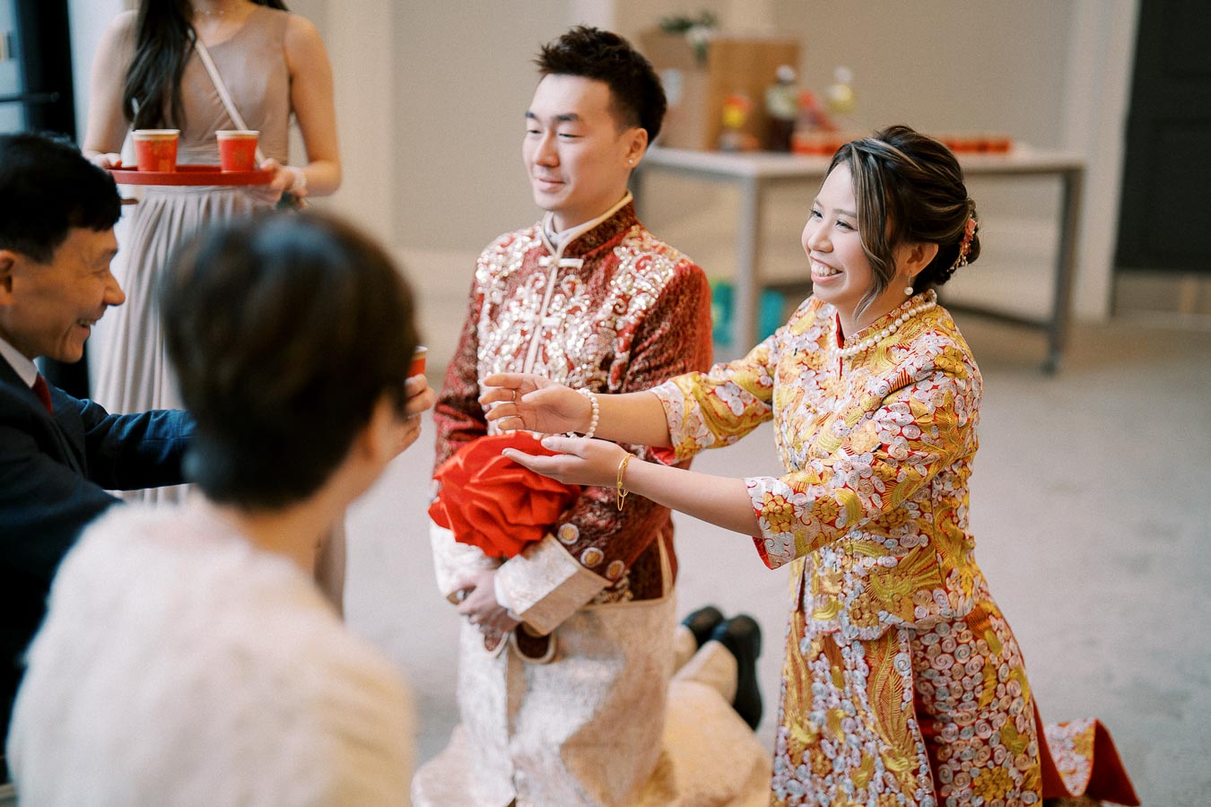 Chinese wedding tea ceremony with a couple in traditional attire presenting tea to elders, smiling bride in yellow and red embroidered dress, and groom in red and white brocade robe, signifying respect and family unity.