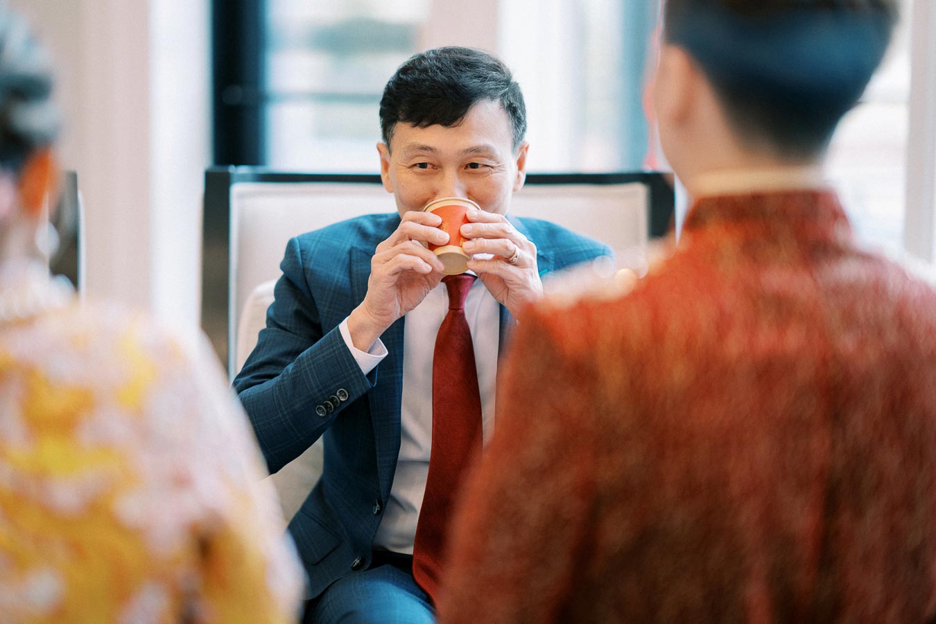 Man in a blue suit sipping from a red cup during a formal gathering, with colorful clothing in the foreground, in a bright indoor setting.