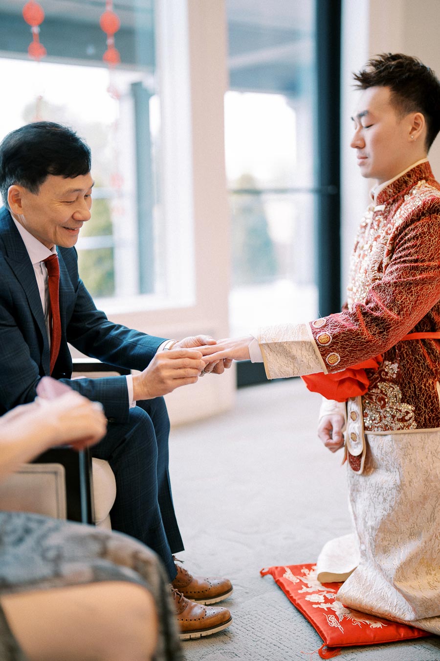 Man in traditional attire kneeling and receiving a ring from another man in a suit during a ceremony, with festive red decorations in the background.