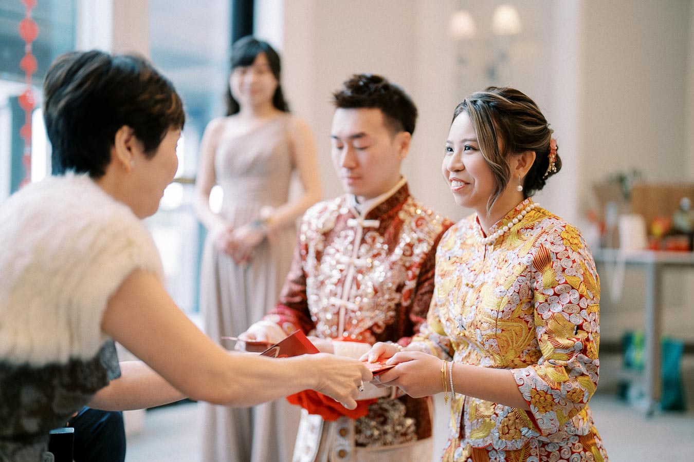 A couple in traditional Chinese wedding attire participates in a tea ceremony, exchanging red envelopes with a seated guest in a modern setting.