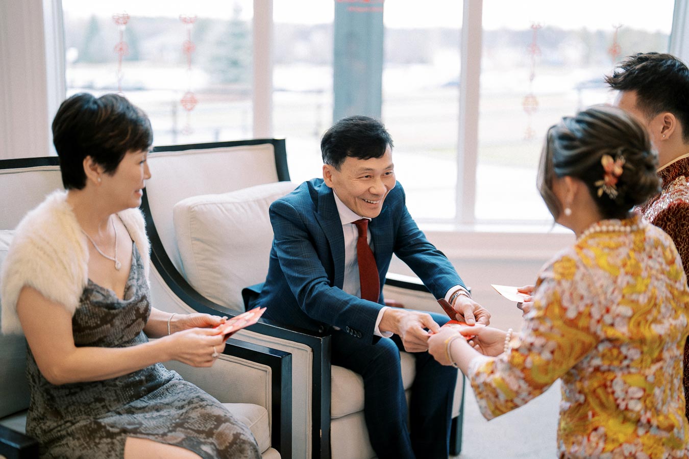 A group of people engaging in a traditional ceremony, exchanging red envelopes in a brightly lit room. The setting suggests a cultural event celebrating good fortune and prosperity.