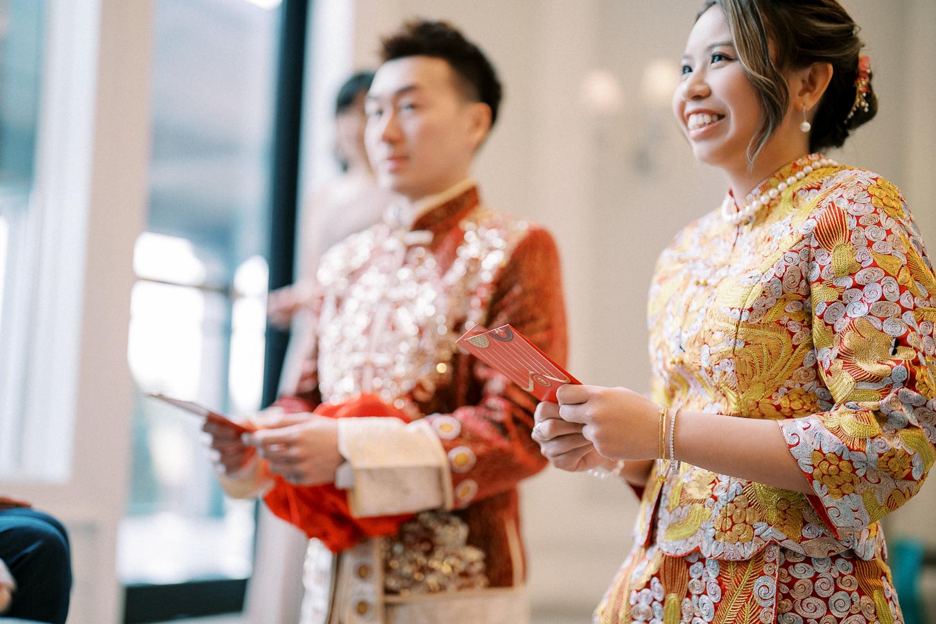 Couple in traditional Asian wedding attire holding red envelopes during tea ceremony indoors.