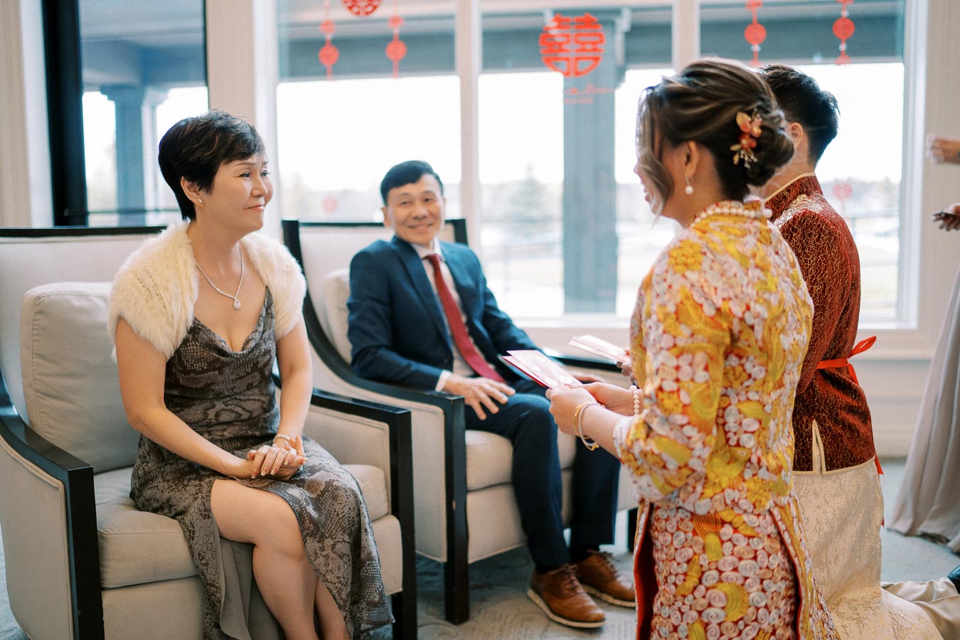 A traditional family gathering featuring a woman smiling while seated in an elegant dress, with a man in a suit, as two individuals in colorful, cultural attire present gifts in a room adorned with festive decorations.