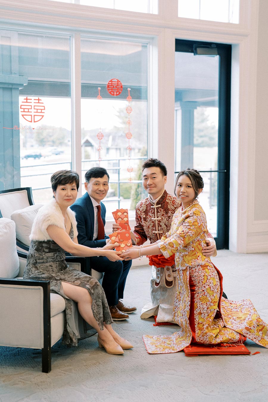 A couple in traditional Chinese attire exchanging red envelopes with family members during a tea ceremony inside a bright room with decorative window decals.