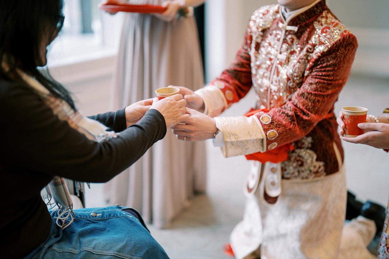 Person in traditional embroidered attire serving tea to a seated individual, symbolizing a cultural ceremony or ritual, with a focus on the ornate details of the clothing and elegance of the interaction.