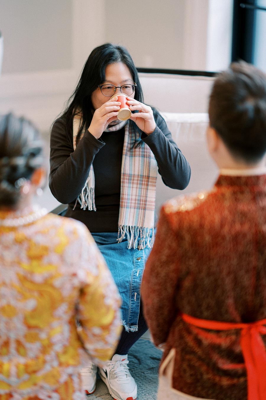 A person wearing glasses and a scarf enjoying a warm drink from a cup, surrounded by others in traditional attire, in an indoor setting.