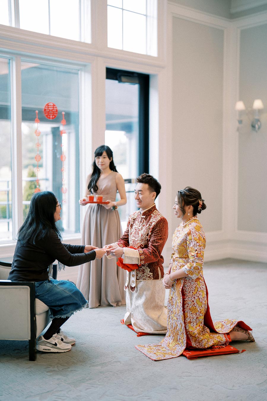 A couple in traditional attire participating in a tea ceremony, with a woman serving tea on a tray, inside a bright room with festive decorations.