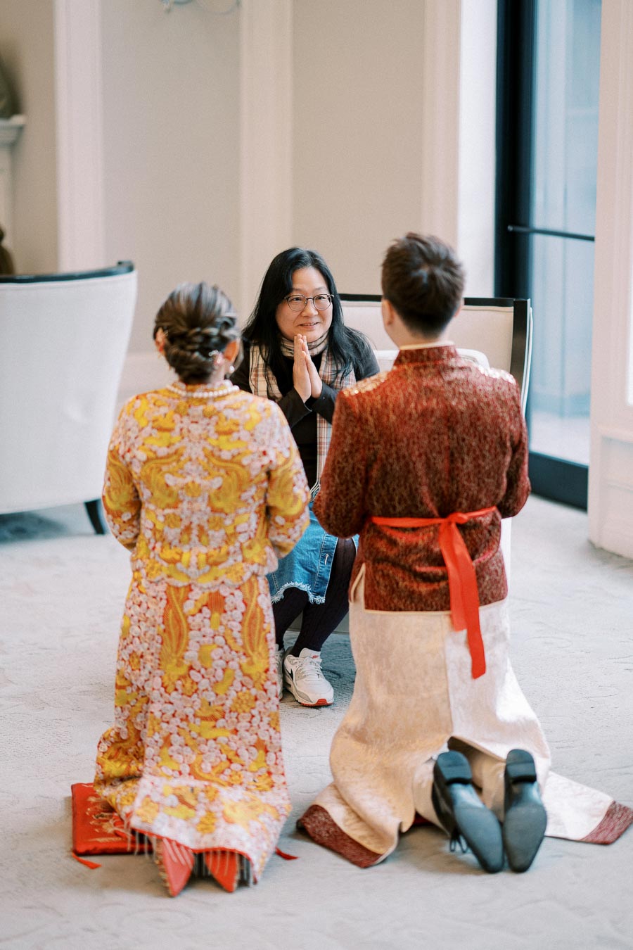 A couple dressed in traditional Asian attire kneeling on a carpeted floor, facing a seated woman with hands in a prayer gesture, inside a bright room with large windows.