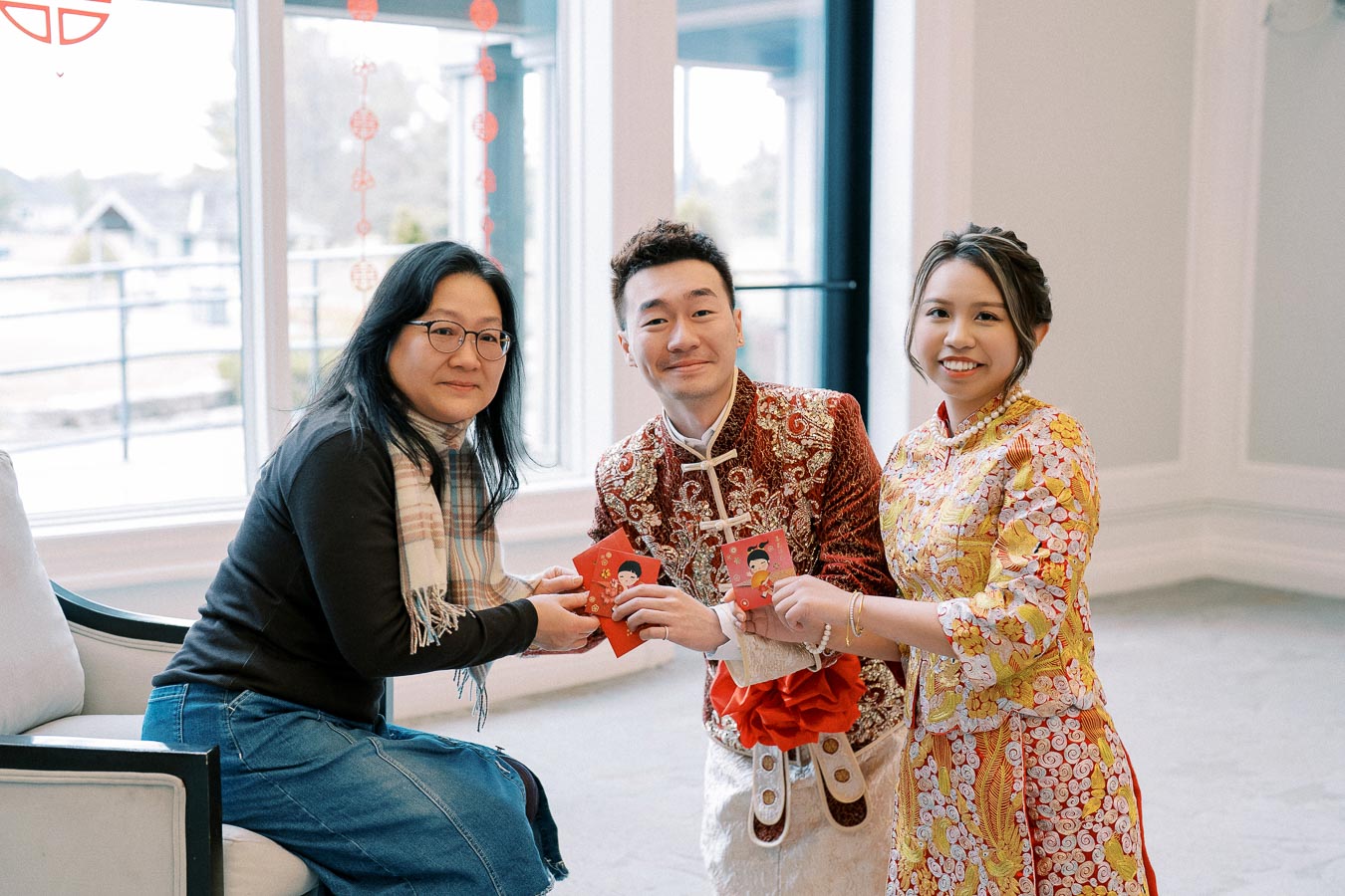 A woman in a scarf and glasses sitting on a chair handing red envelopes to a man and woman dressed in traditional Chinese attire, celebrating Lunar New Year in a bright room decorated with festive ornaments.