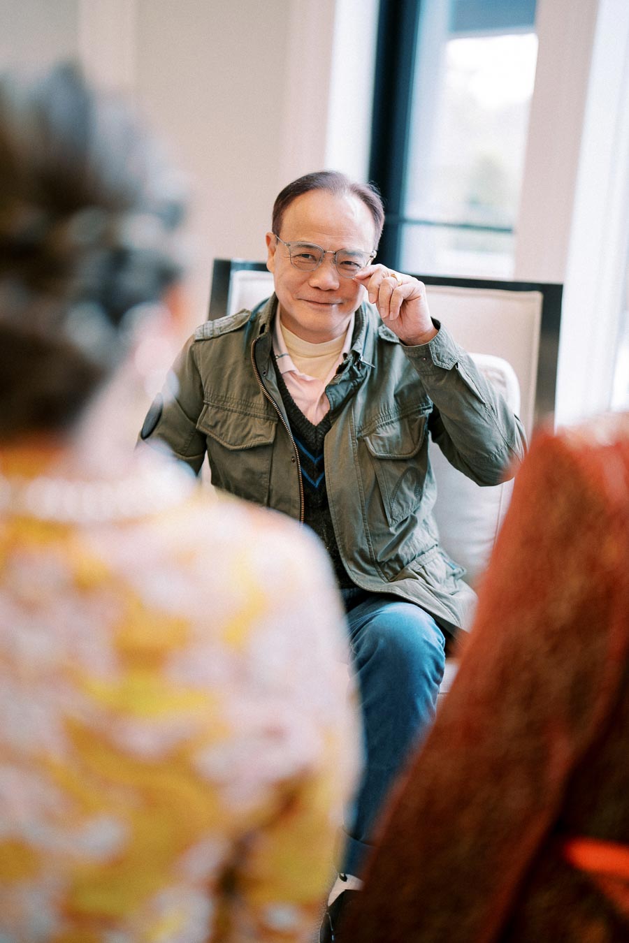 Man in a green jacket smiling warmly while adjusting glasses, engaged in a conversation with two blurred individuals in the foreground in a brightly lit room.