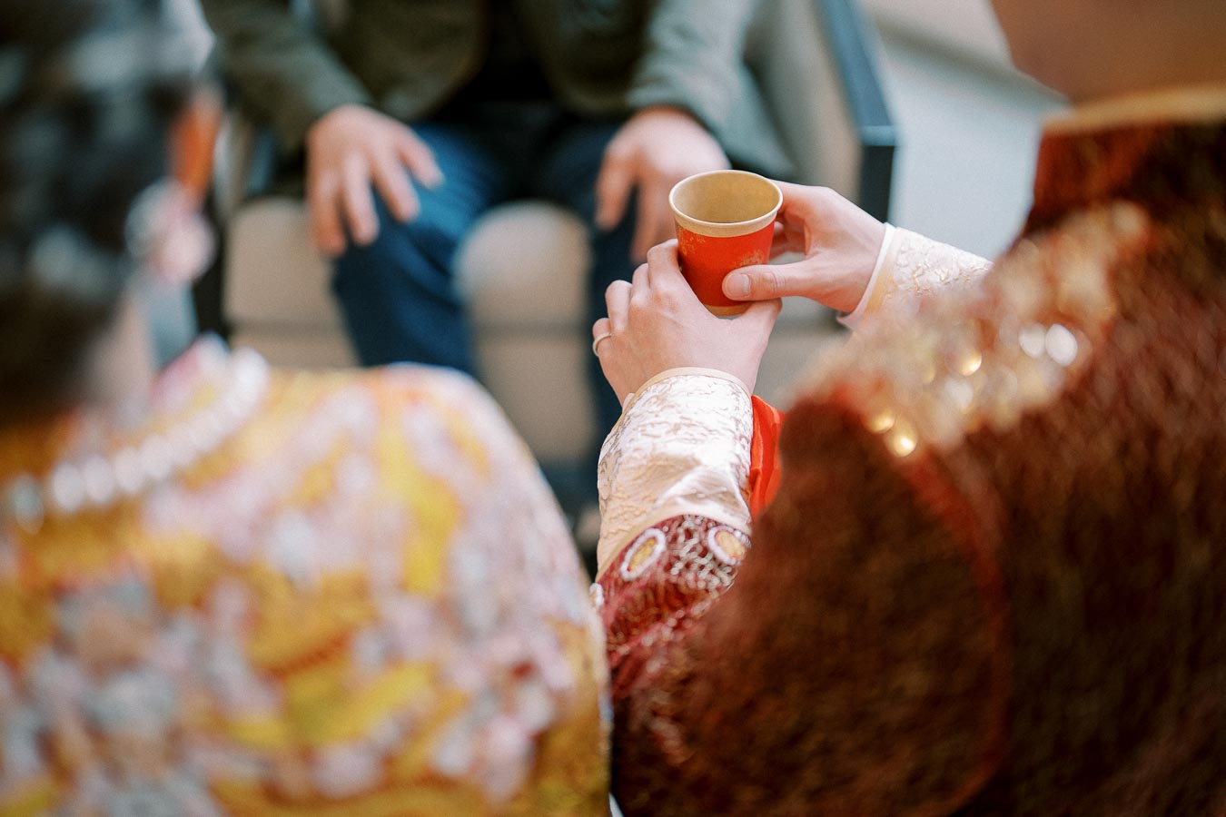 Traditional tea ceremony, close-up of hands holding red cup, participants in ornate garments, seated person in background.