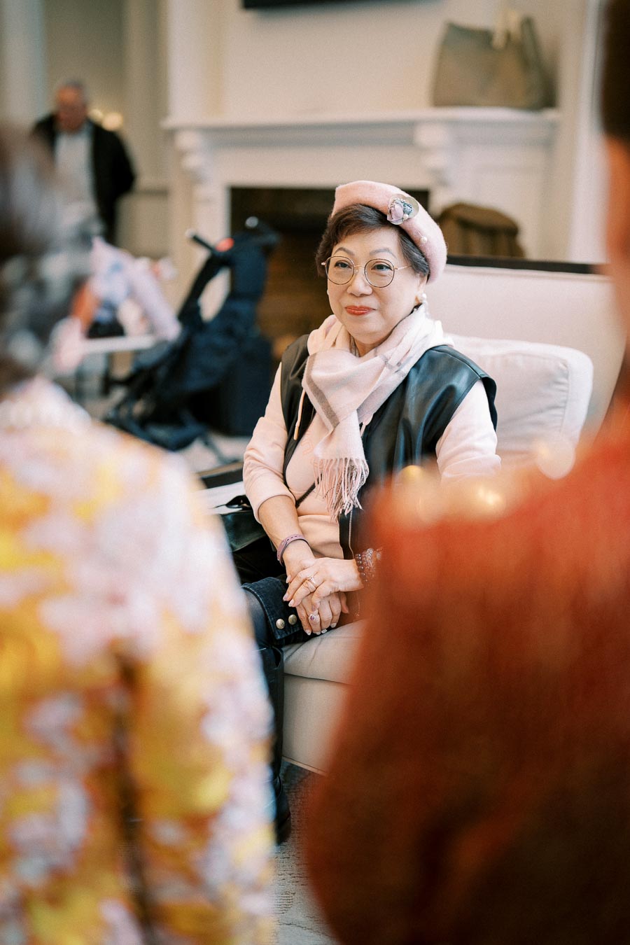 Senior woman wearing a pink hat and scarf sitting comfortably in a living room setting, engaging in a conversation.