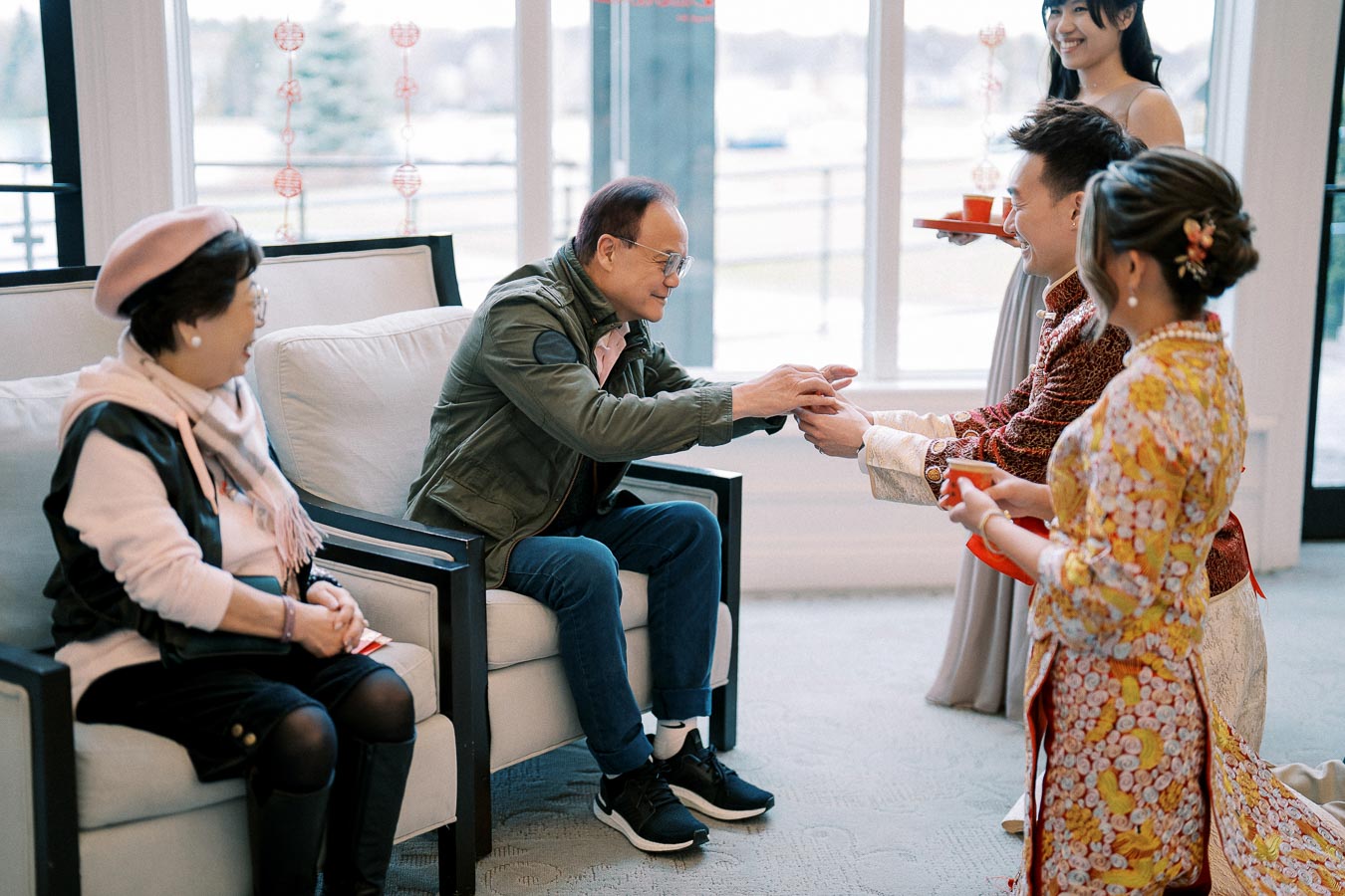 A group of people participating in a traditional tea ceremony indoors. The individuals are dressed in elegant attire, with one person handing tea to an older couple seated in chairs. Natural light streams through large windows in the background, creating a warm and inviting atmosphere.