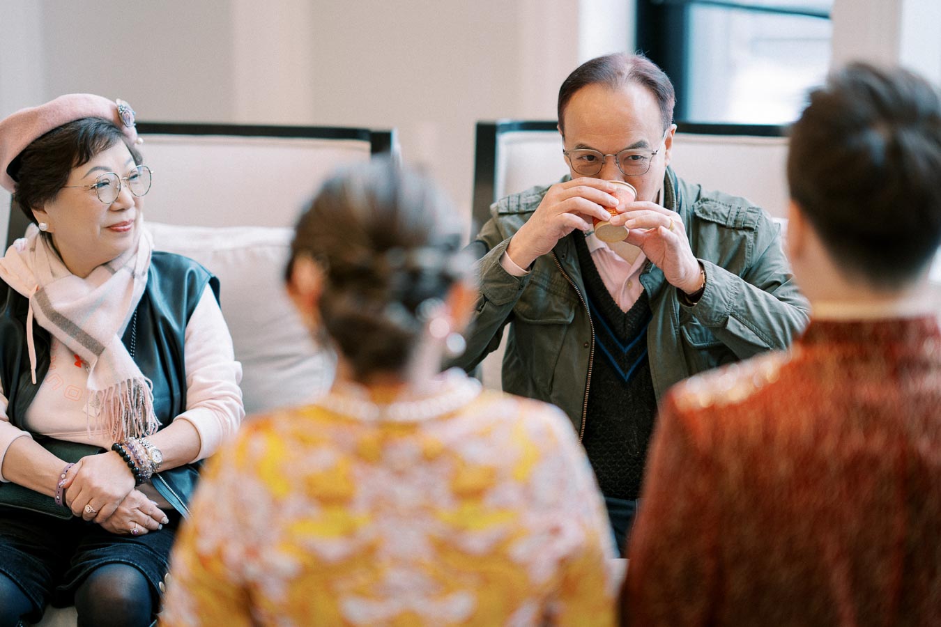 Group of people enjoying a casual gathering indoors, with an older man sipping a drink and a woman wearing glasses and a beret sitting nearby.
