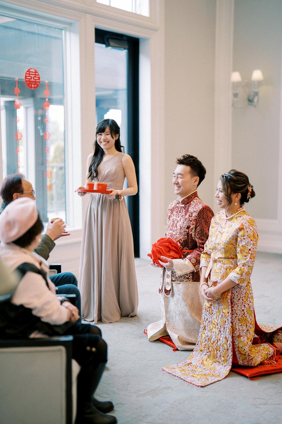 Traditional Chinese wedding tea ceremony with a couple dressed in elaborate red and yellow attire kneeling before seated family members, while a woman in an elegant gown presents tea.