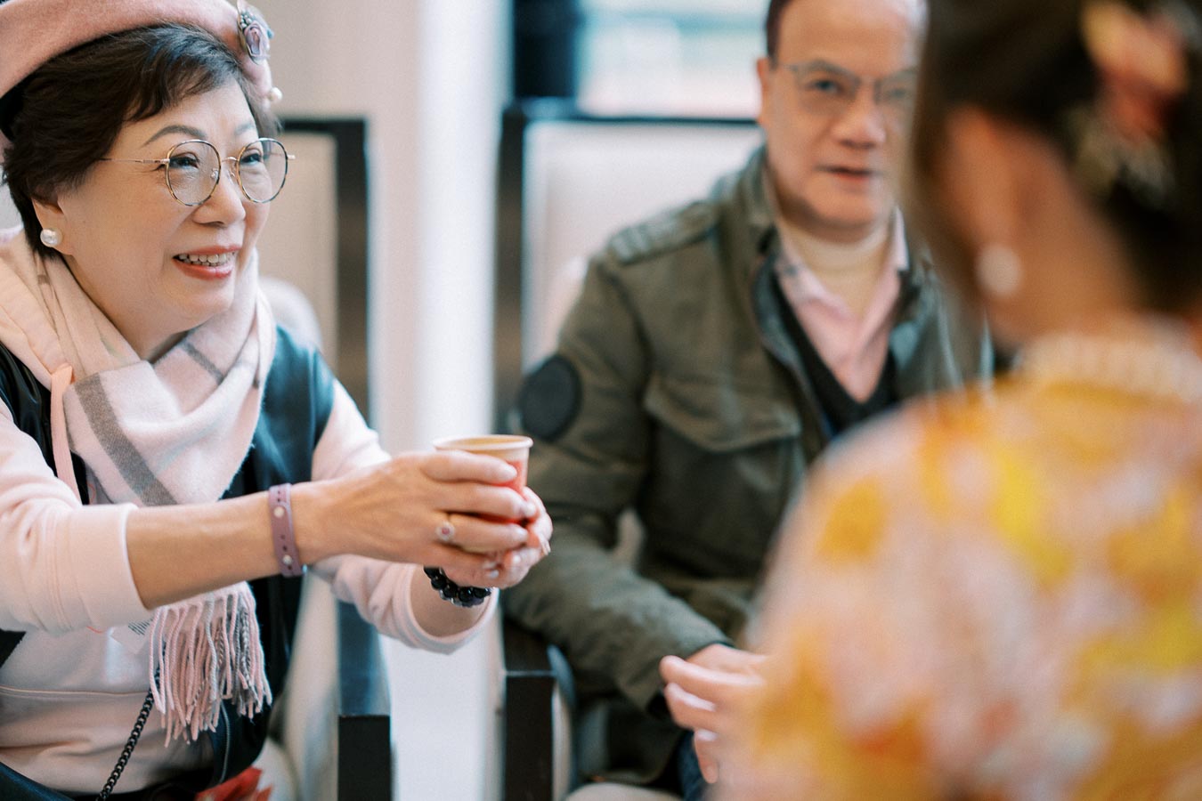 Elderly woman smiling and offering a cup during a social gathering, with a man sitting beside her, illustrating warmth and hospitality.