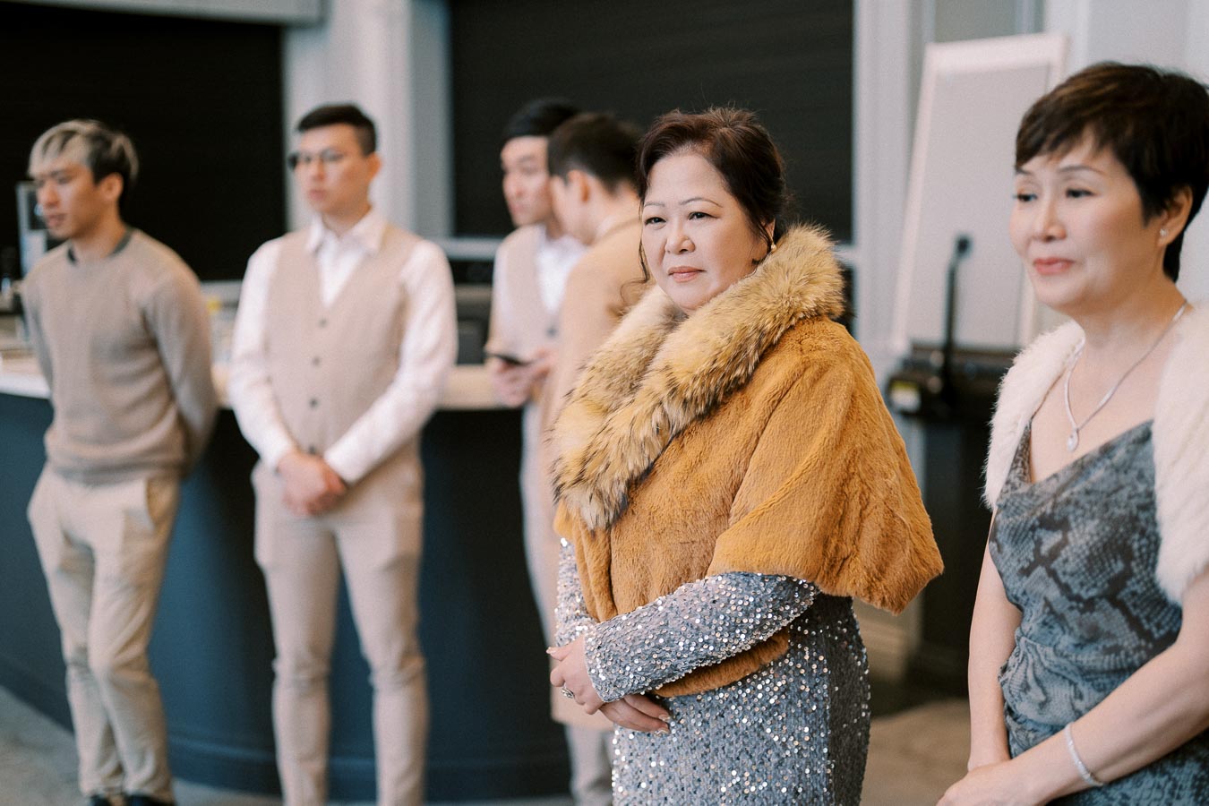 A group of elegantly dressed people at a formal gathering, with two women in the foreground wearing fur-trimmed shawls and sparkling dresses, while three men stand in the background wearing beige outfits.