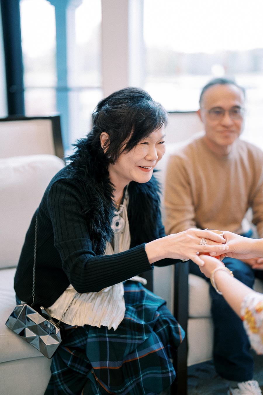 Smiling woman sitting on a couch, holding hands with another person, while a man sits nearby in a cozy, well-lit room.