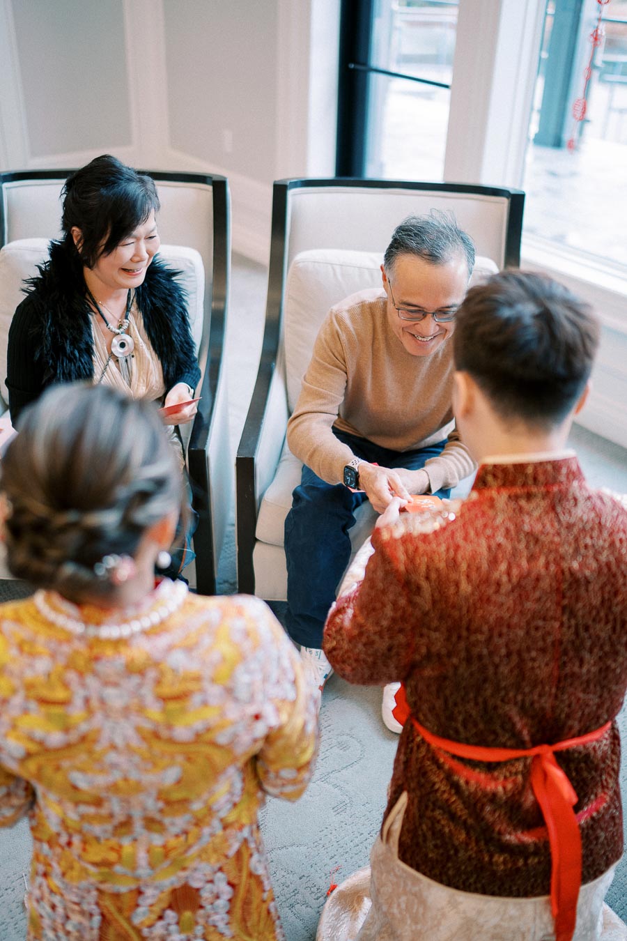 Elderly couple seated smiling during a traditional tea ceremony with two people in colorful attire presenting tea, symbolizing respect and celebration.