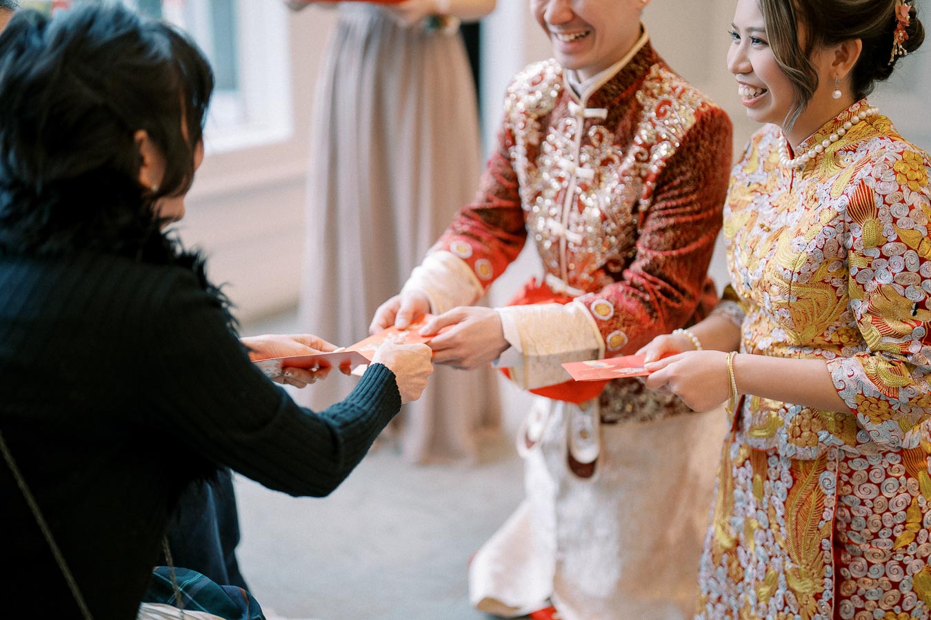Couple in traditional Asian attire smiling while exchanging red envelopes during a cultural ceremony indoors.