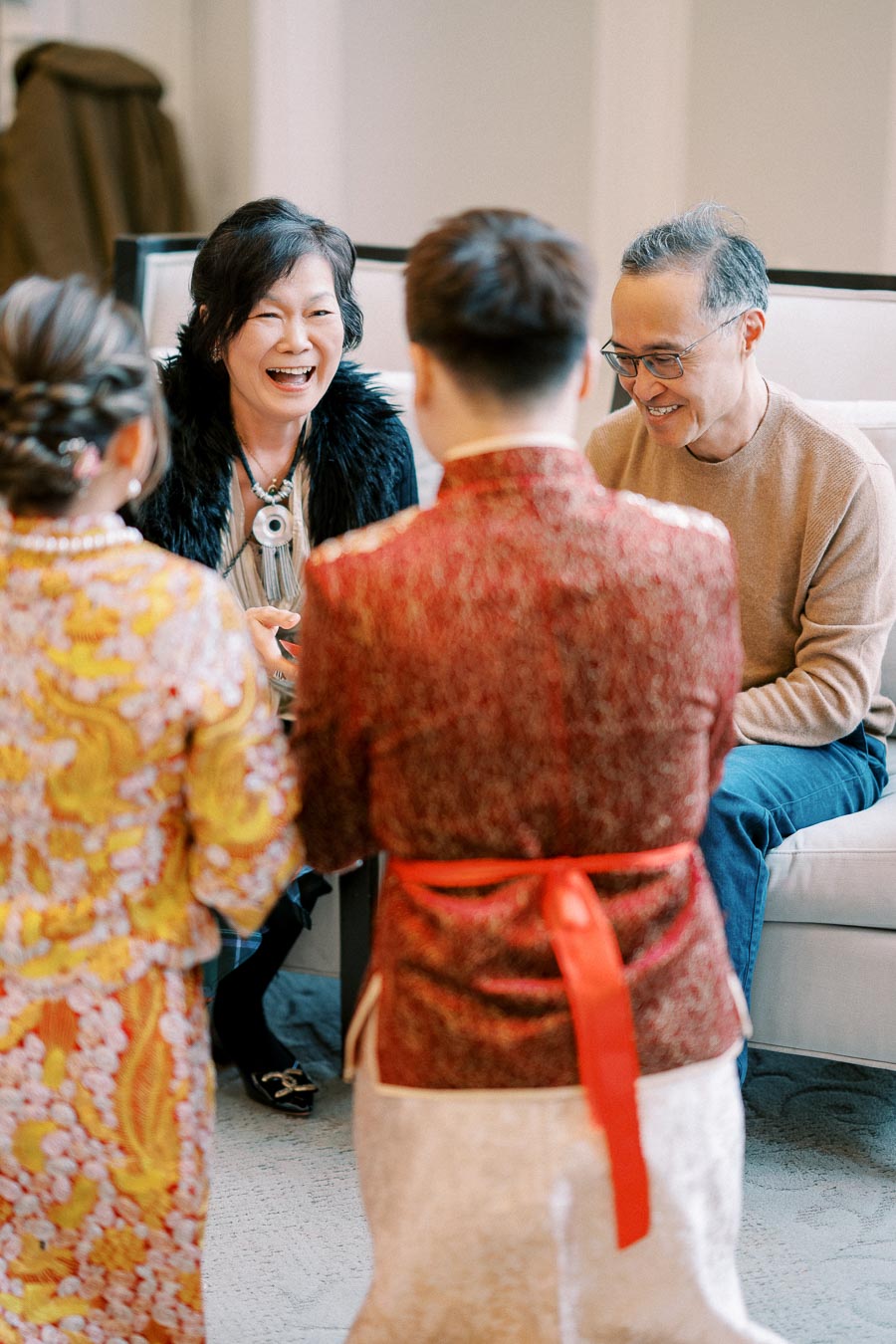 A joyful group of four people, two standing in traditional attire facing a seated couple, who are smiling and engaging in a warm, happy conversation indoors.