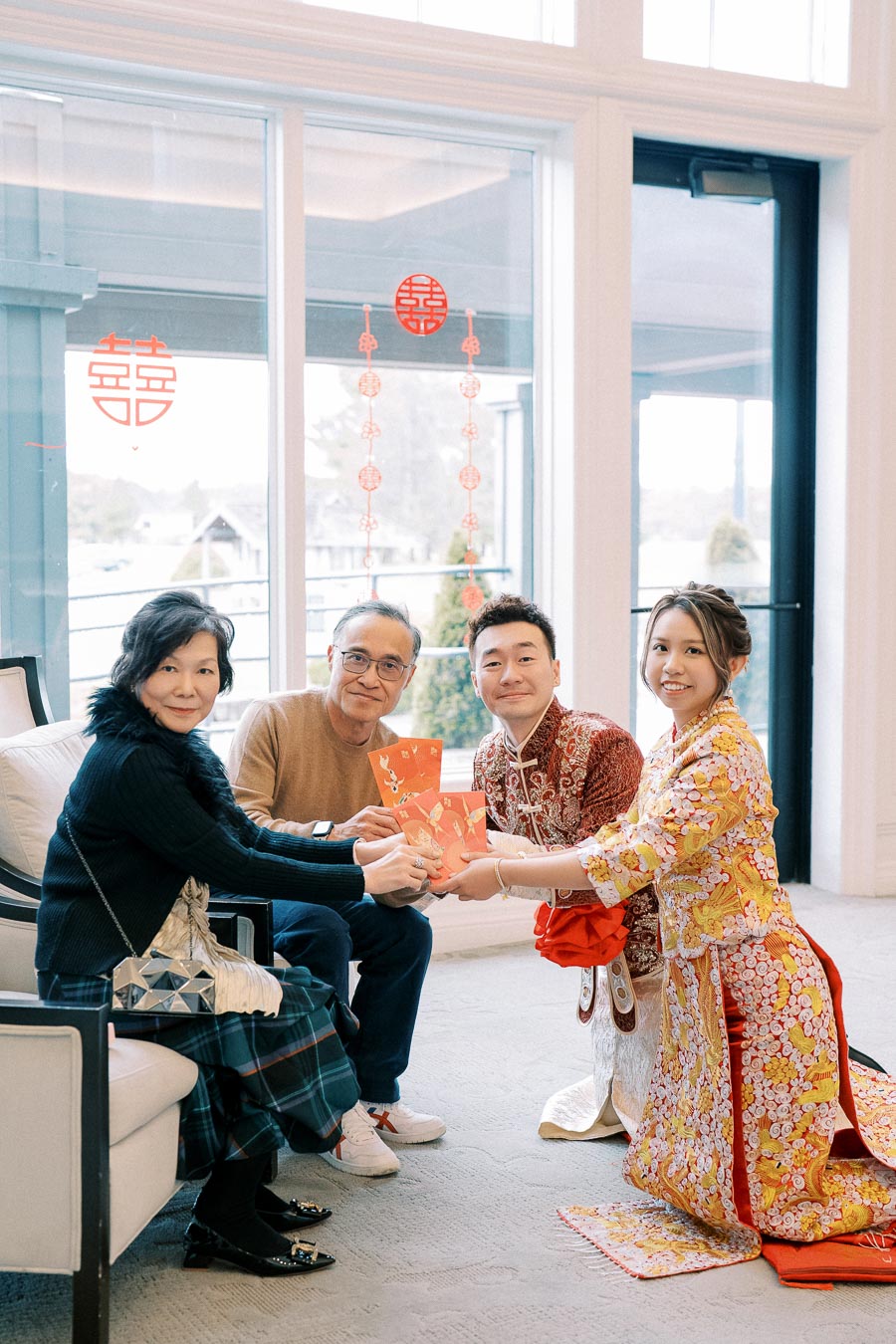A family gathered celebrating Chinese New Year, exchanging red envelopes while dressed in traditional attire, with festive decorations in the background.