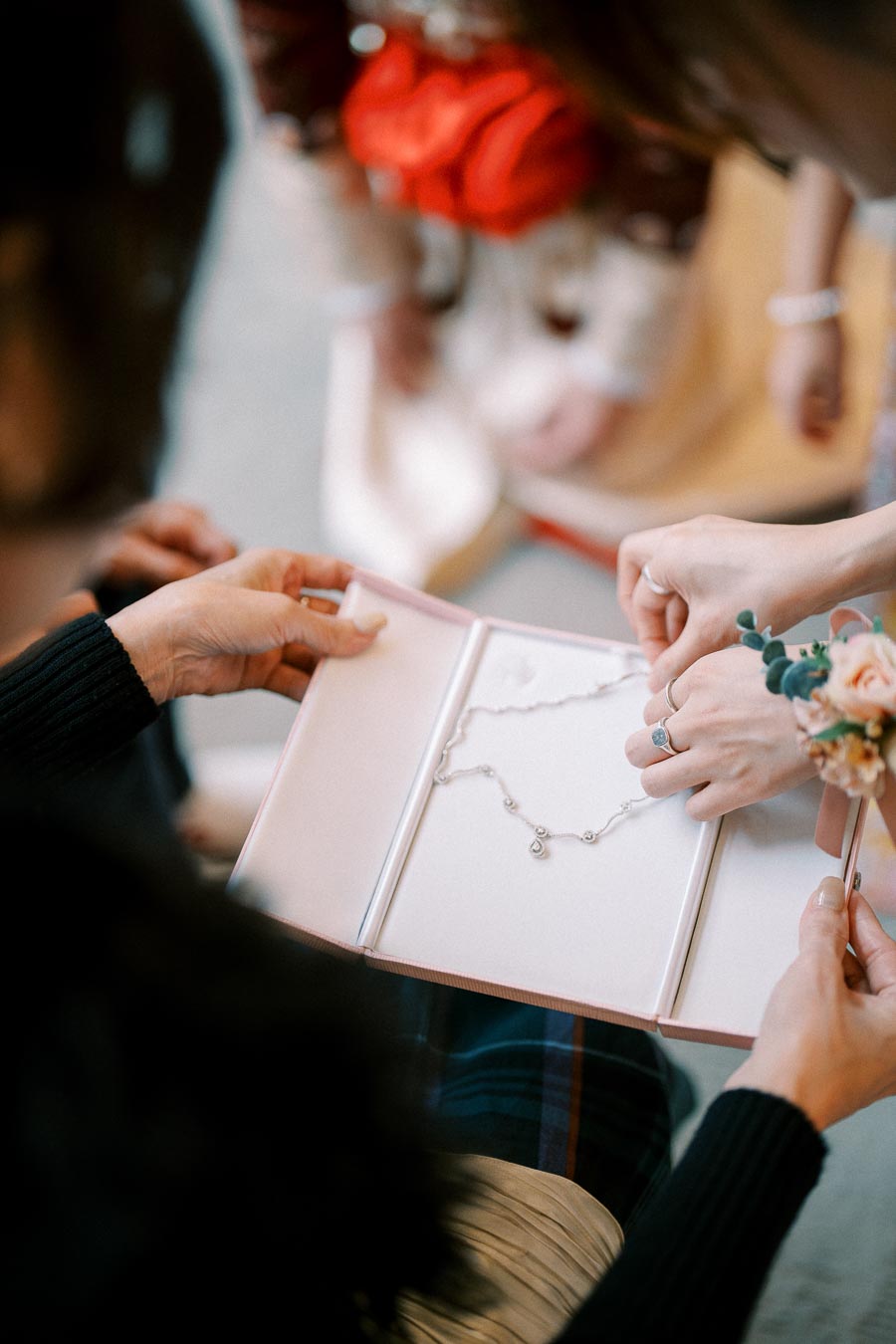 Hands opening a pink jewelry box containing a delicate silver necklace with a charm, surrounded by soft focus and floral elements, creating a sense of elegance and celebration.