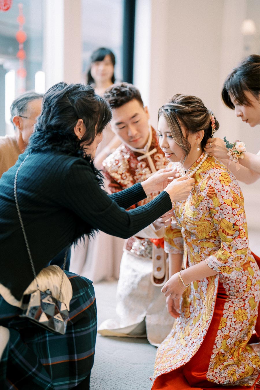 A bride and groom in traditional Asian wedding attire, surrounded by family members helping with jewelry, during a heartfelt wedding ceremony.