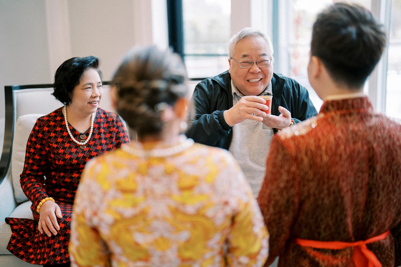 Elderly man holding a cup and smiling, seated beside a woman in a red patterned dress, engaging in a traditional tea ceremony with two younger individuals in formal attire.