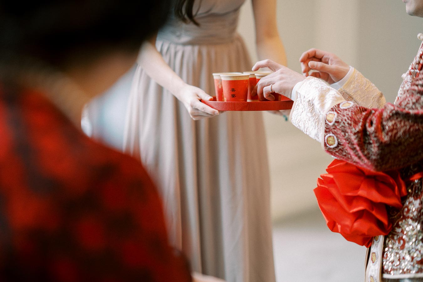 A person in a traditional red and gold embroidered outfit receives a tray of red cups during a ceremonial event, symbolizing cultural celebration and hospitality.