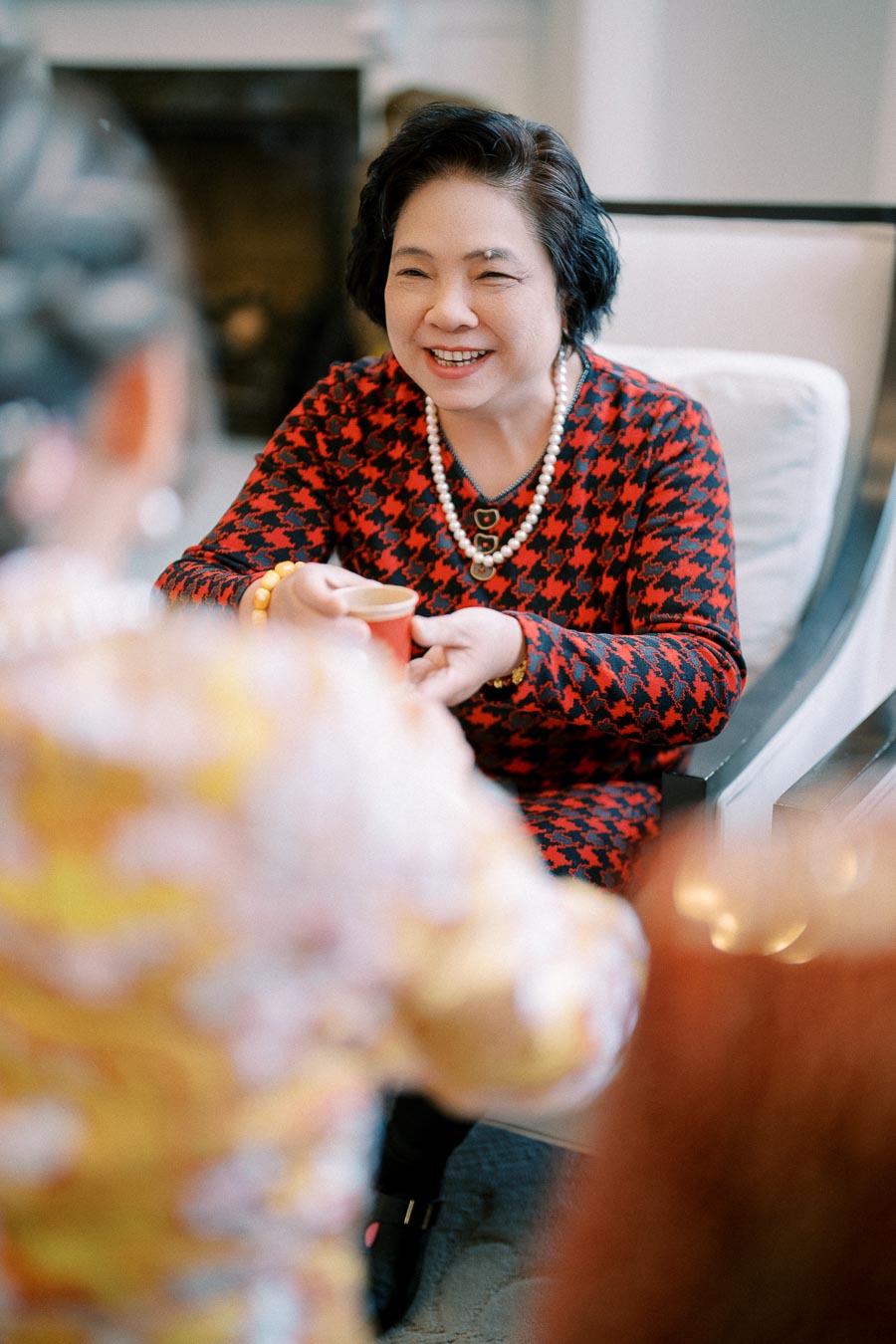 A smiling woman wearing a red and black houndstooth dress and pearl necklace receives a cup in a warmly lit room.