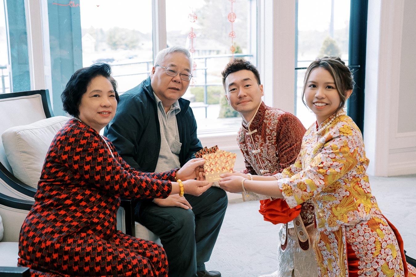 A family celebrating Lunar New Year indoors, exchanging red envelopes. The older couple sits on a sofa wearing festive clothing, while the younger couple dressed in traditional attire stands, smiling and presenting gifts. Bright daylight filters through large windows adorned with festive decorations, creating a warm and joyful atmosphere.