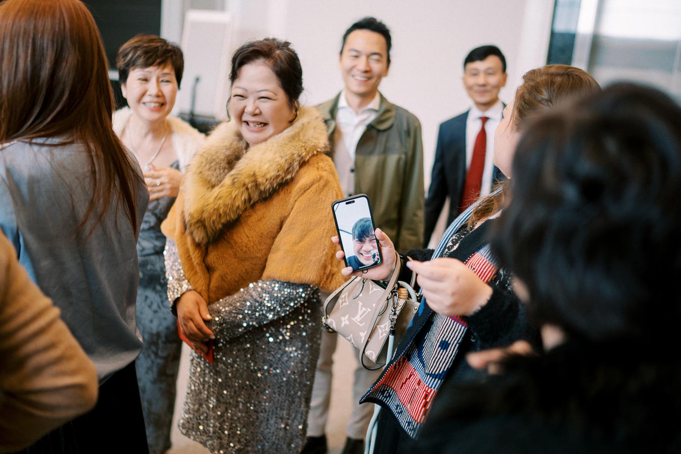 A group of people smiling and engaging in a lively conversation at an indoor gathering, with a woman in a fur-collared coat and a sequin dress showing a video call on her smartphone, creating a joyful and social atmosphere.