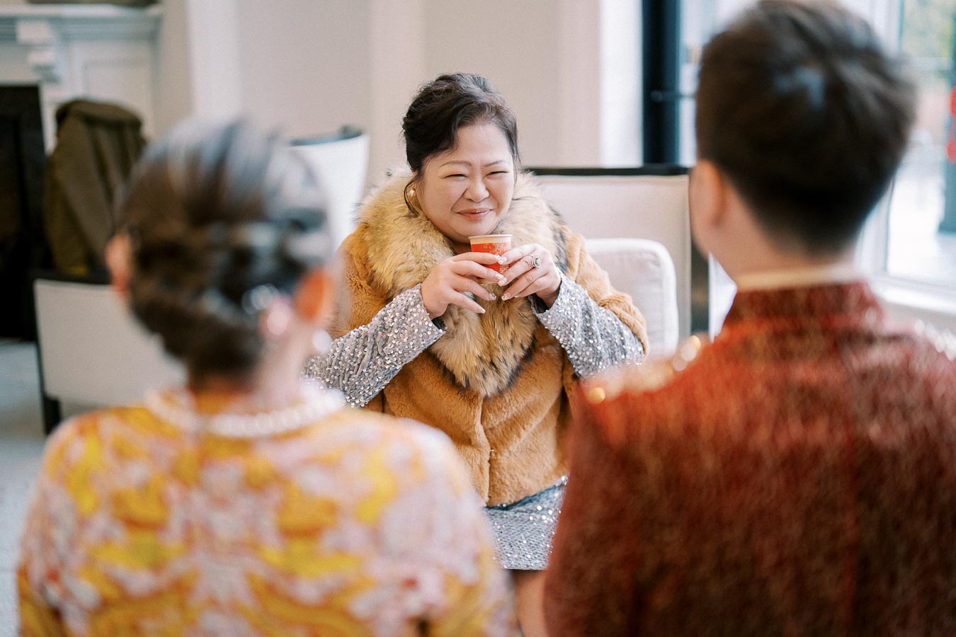 Smiling woman in luxurious fur-trimmed coat enjoying tea during a formal gathering, facing two people dressed in traditional attire.