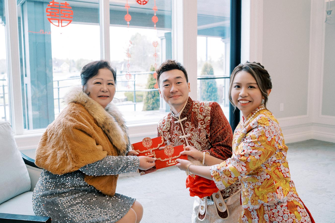 A smiling family exchanging red envelopes during a Chinese celebration, dressed in traditional attire with festive decorations in the background.