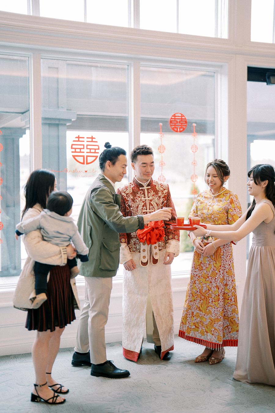 A family participating in a traditional Chinese tea ceremony, with the couple dressed in ornate attire, surrounded by decorations symbolizing happiness, in a bright room with large windows.
