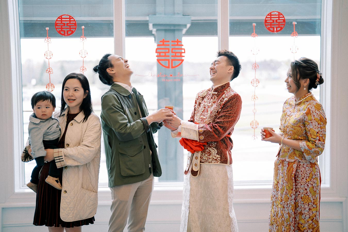 A joyful group at a Chinese wedding celebration, featuring a couple in traditional attire exchanging tea, surrounded by friends and family with festive decorations in the background.