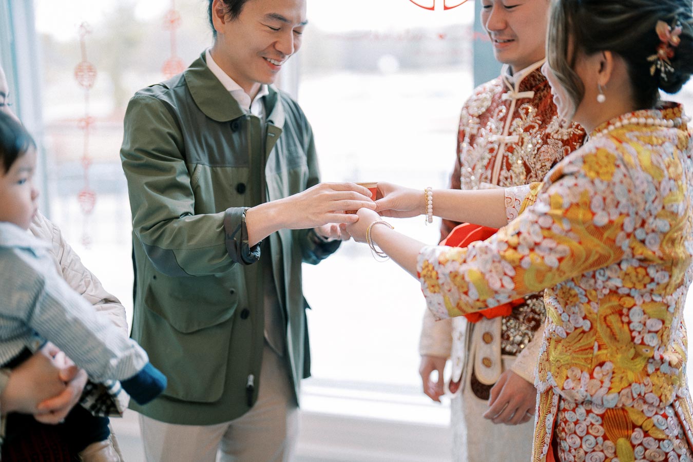 A couple in traditional Chinese wedding attire performs an exchange ceremony, surrounded by family, highlighting cultural customs and joyful celebration.