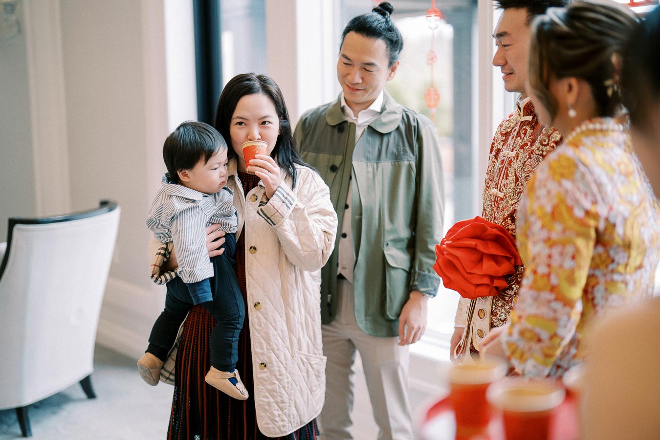 A group of people celebrating a festive occasion; a woman holding a baby and drinking from a red cup, joined by others in colorful attire.