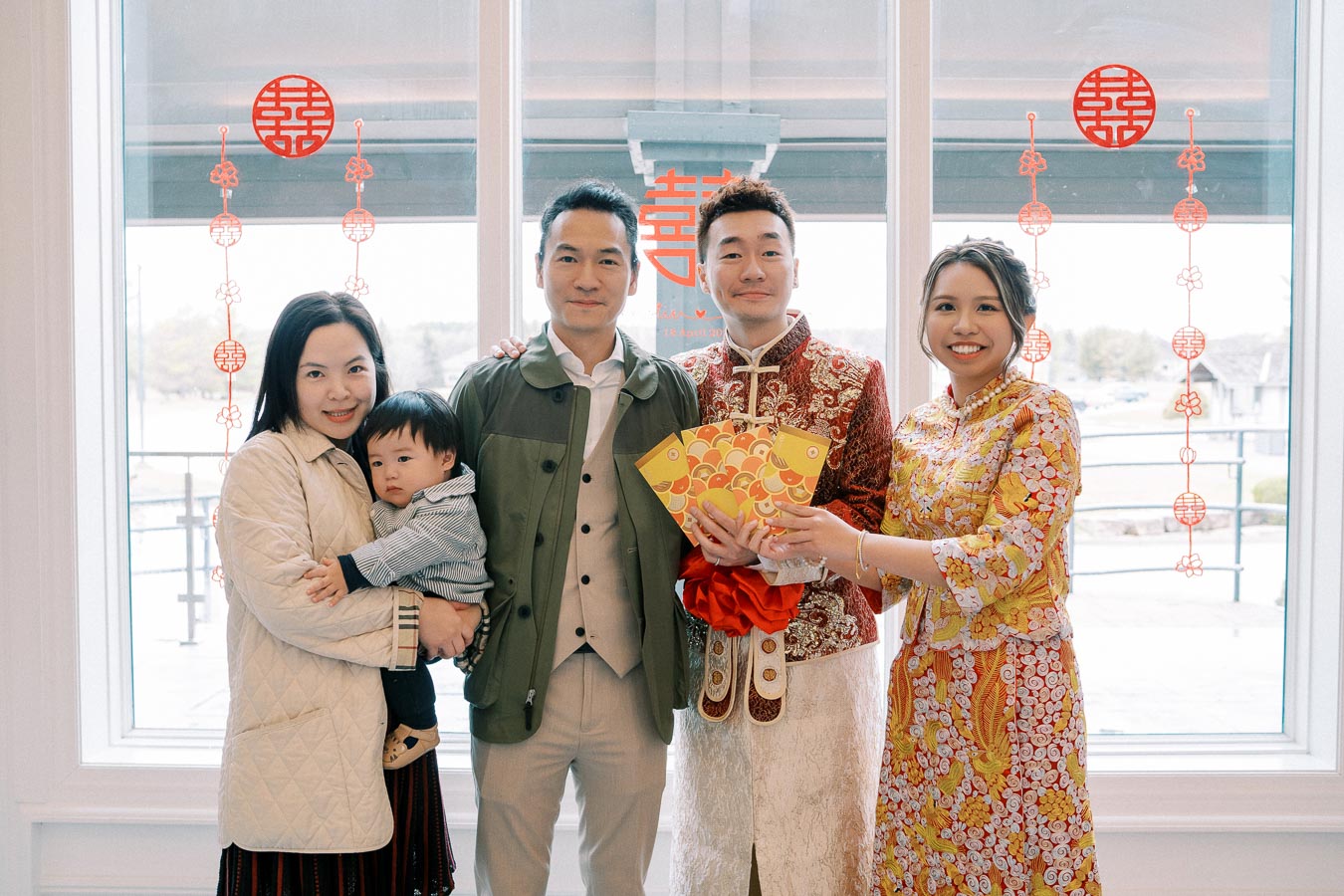 Family celebrating a traditional event indoors, with red and gold decorations symbolizing good fortune. A couple wears ornate traditional outfits, holding colorful envelopes, surrounded by family members, including a child. Red Chinese characters adorn the windows in the background.