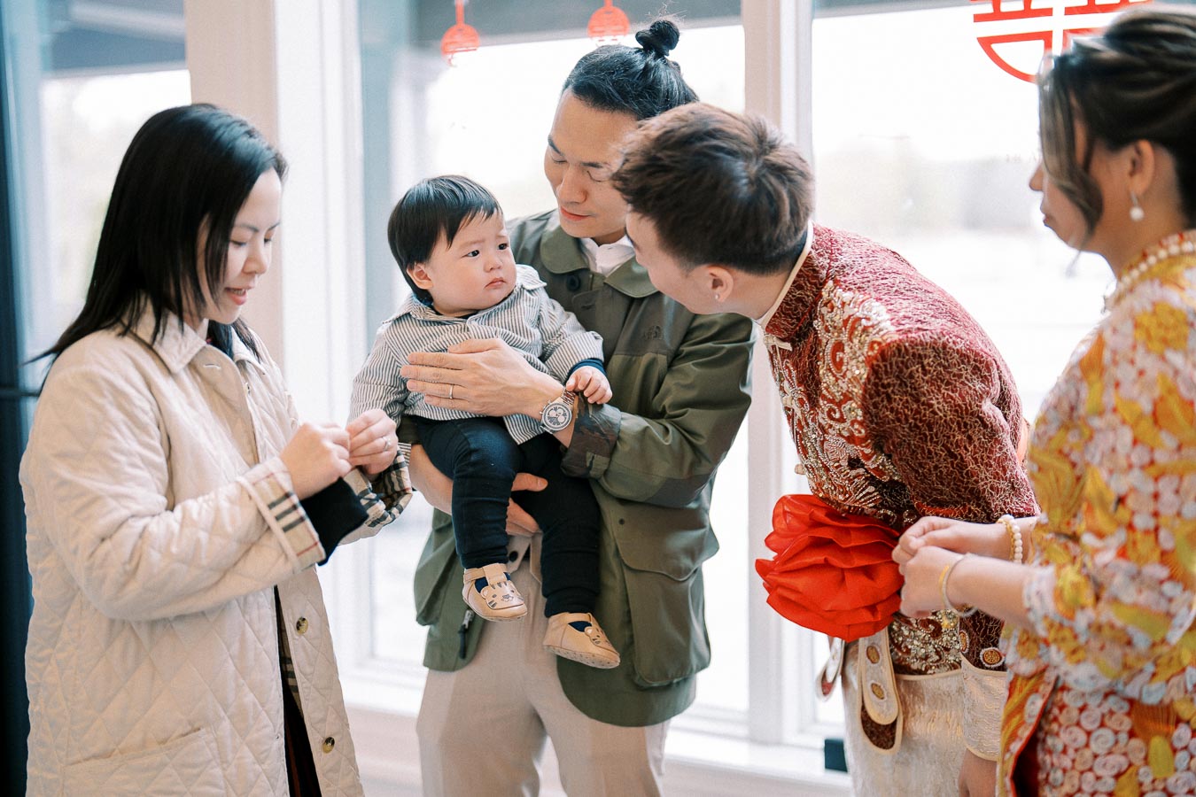 A family gathered during a traditional celebration, featuring a child being held by a parent. Another adult in ornate attire leans in, while others exchange smiles, in a warmly lit indoor setting.