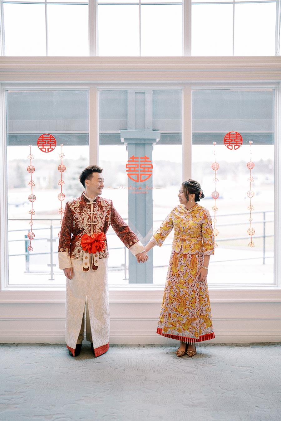 A couple in traditional Chinese attire, holding hands and smiling at each other, with red double happiness decorations on the window behind them.