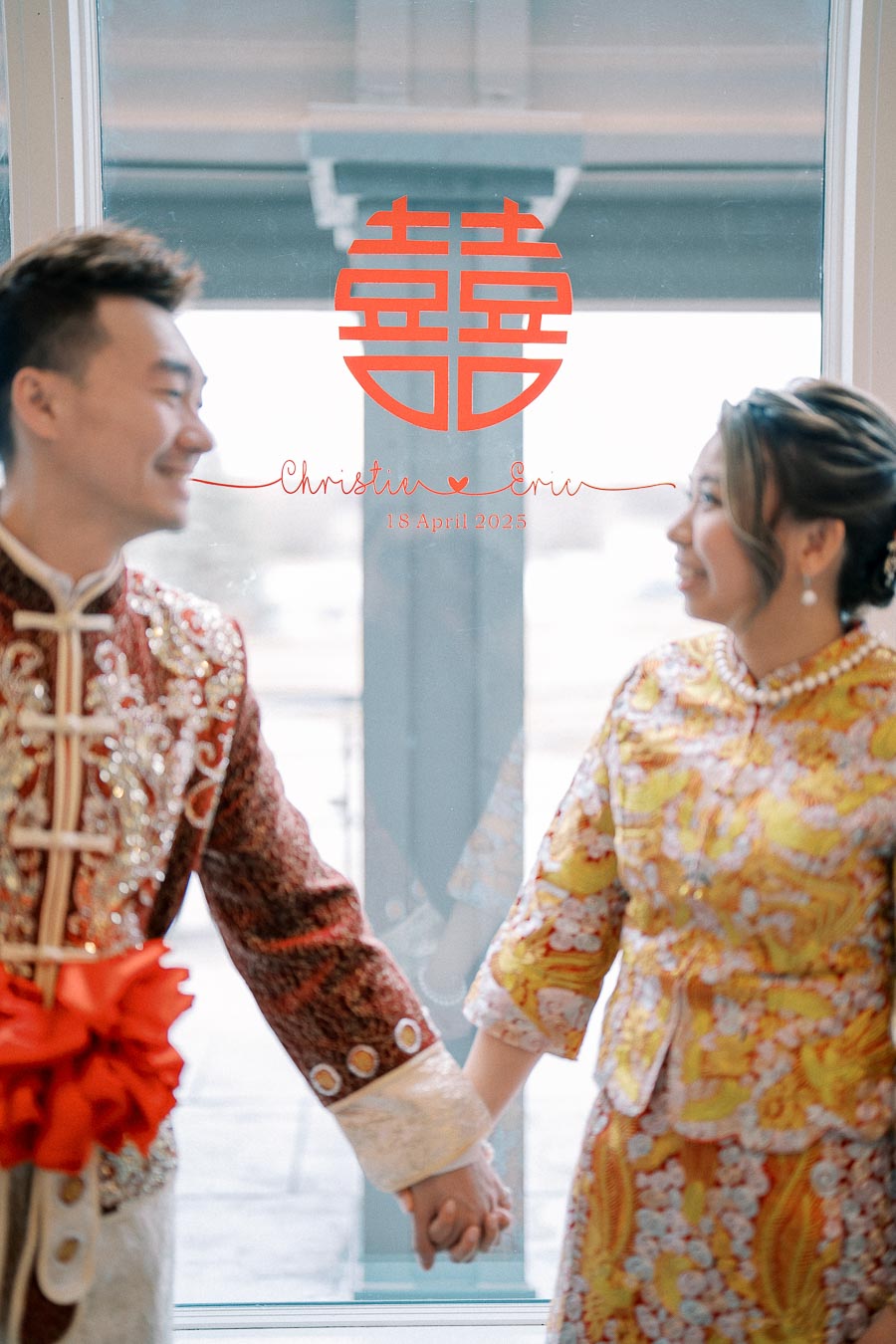 Elegant couple in traditional Chinese wedding attire holding hands, with a double happiness symbol and personalized wedding details on a window in the background.