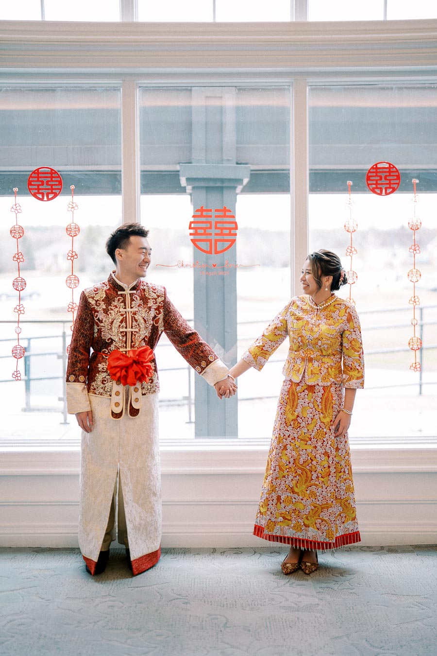A couple in elegant traditional Chinese wedding attire holding hands, standing before a window with symbolic red Chinese characters and decorations.