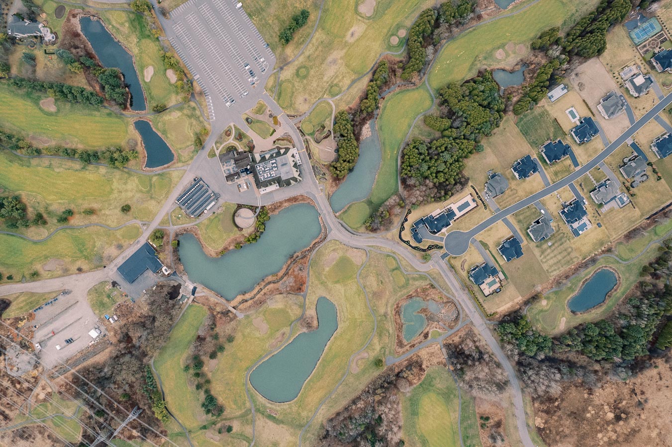 Aerial view of a golf course with green fairways and sand bunkers, surrounded by residential homes and parking area, showcasing a landscape of trees and small ponds.