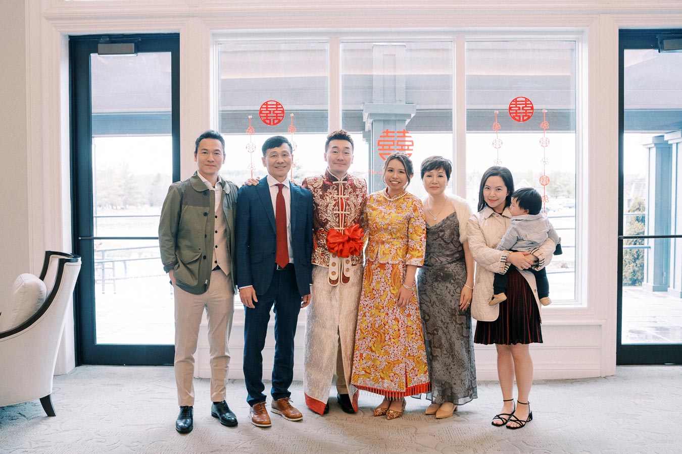 A happy group of people in traditional Chinese attire, standing together indoors by large windows with festive red decorations.