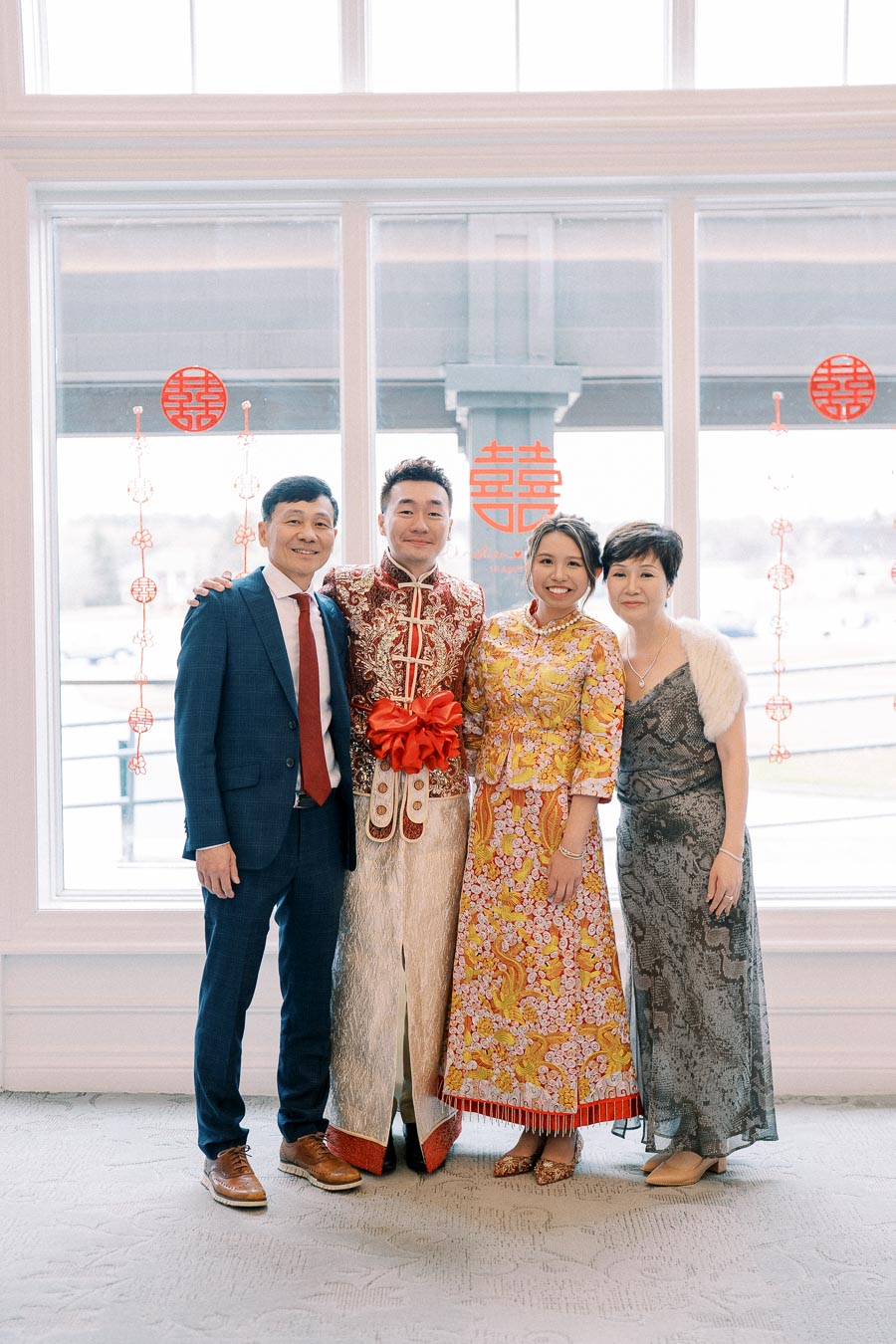 Asian family celebrating a traditional wedding, featuring a couple in ornate festive attire with parents, standing in front of a window decorated with red Chinese symbols for happiness.